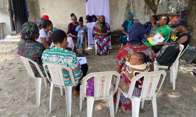 A group of women sit in a circle on plastic chairs outdoors, participating in a community meeting. Several women hold young children, including one woman with a baby wrapped against her back. Some participants hold papers or notebooks, and a banner stands behind the group. The setting appears informal, with a tree and simple building walls in the background. image linking to From silence to connectivity: Nigerian women building digital pathways to justice