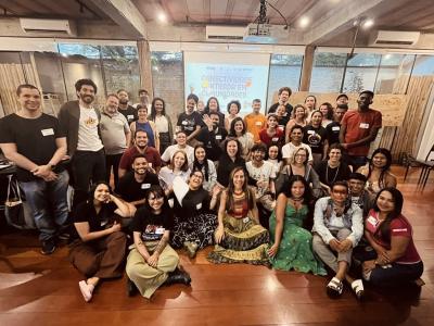 A diversified group of about 50 people smile to a camera in a room. Behind them, a cloth displays a text about connectivity in the communities. In a lively atmosphere. image linking to Updated self-affirmation from the Brazilian National Committee of Community Networks